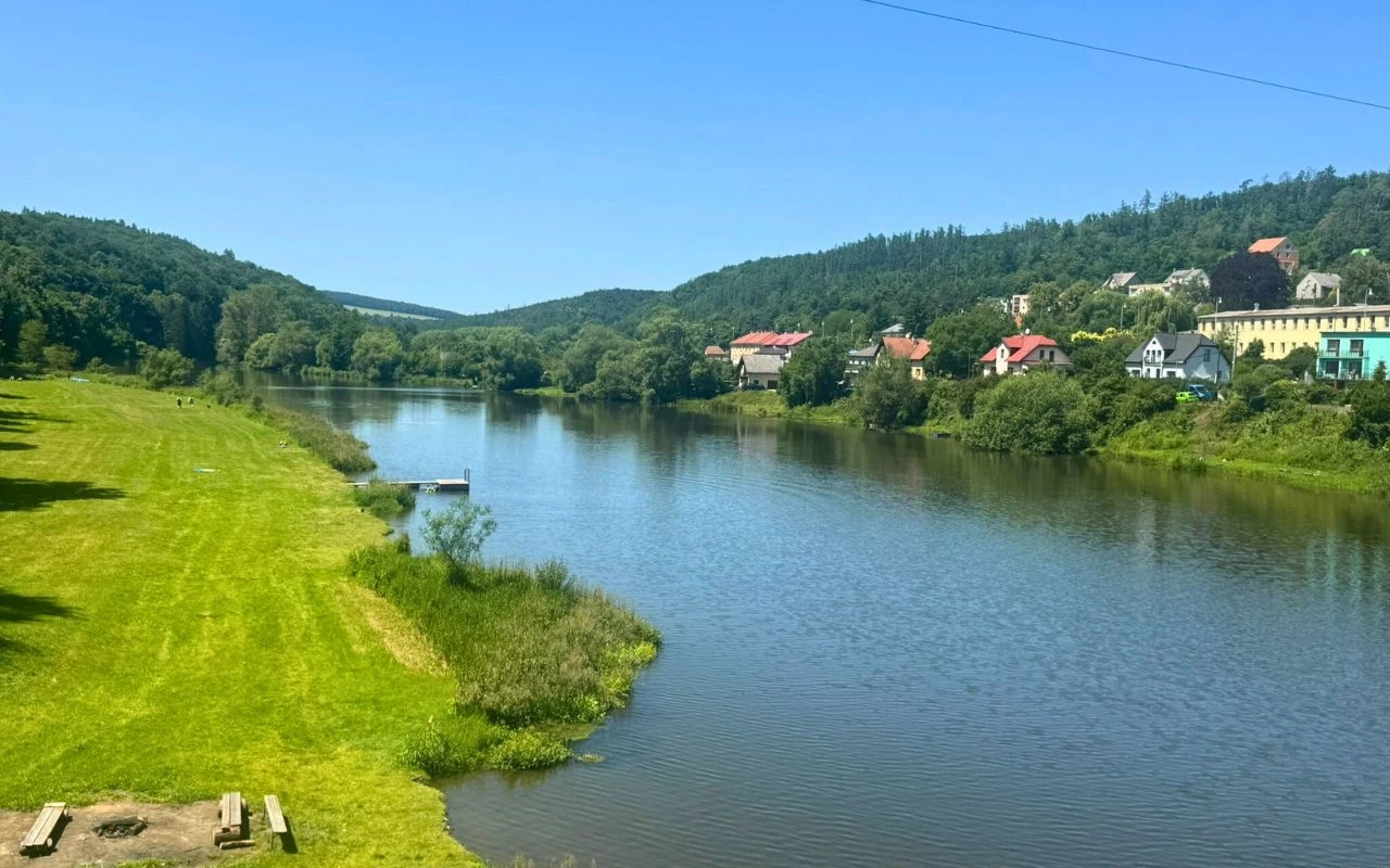 Spokojna rzeka otoczona zielonymi łąkami i wzgórzami, po jednej stronie widoczne są budynki mieszkalne z kolorowymi dachami, a nad wodą znajduje się niewielki pomost.