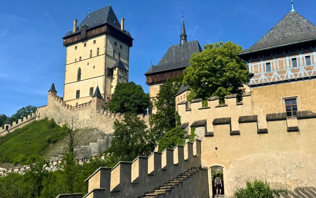 Zamek Karlstejn Historyczny zamek o grubych murach, wysokich wieżach i stromych schodach otoczony zielenią, pod błękitnym niebem, z wyraźnie widocznymi detalami architektonicznymi.