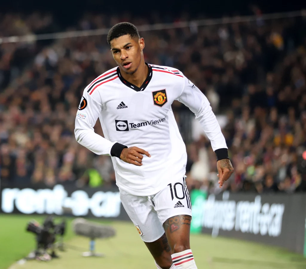 Marcus Rashford Piłkarz w białym stroju Manchesteru United z numerem 10 biegnie po murawie, w tle widoczny tłum kibiców i fragment boiska.