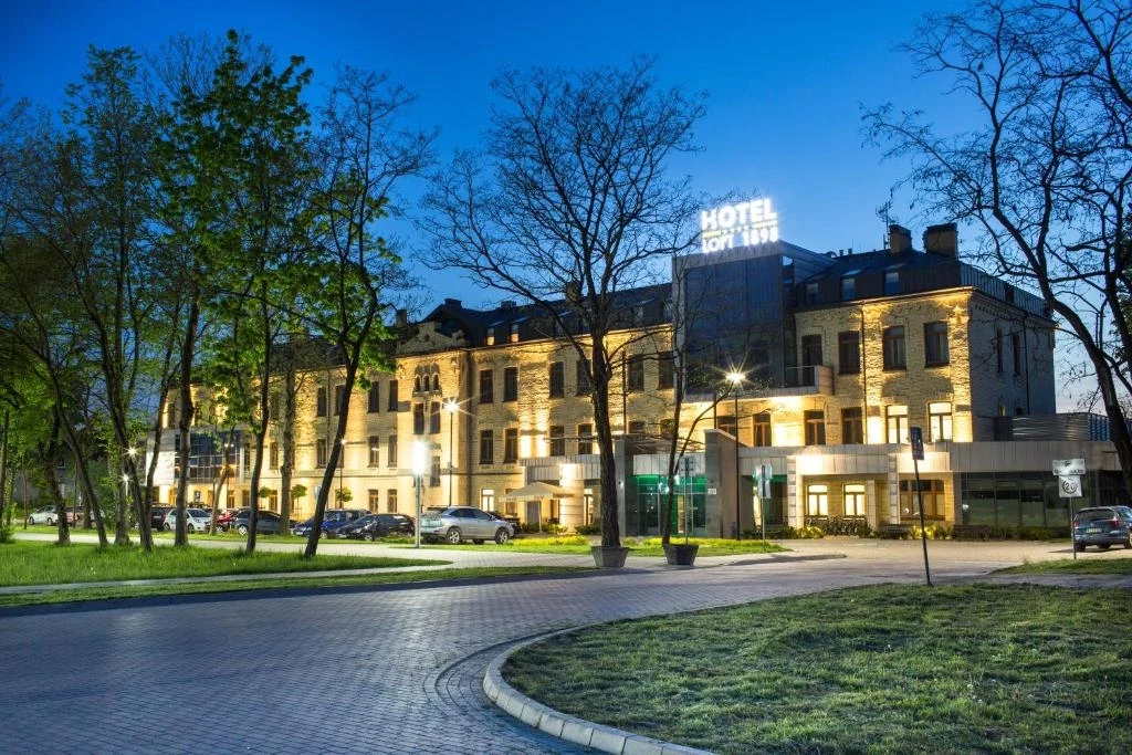 Suwalski hotel LOFT 1898 Duży, elegancki hotel oświetlony światłami, otoczony zadbaną zielenią i drzewami, z widocznym napisem HOTEL na dachu, fasada budynku utrzymana w klasycznym stylu, na parkingu zaparkowane samochody, wieczorna atmosfera podkreślona niebieskim niebem.