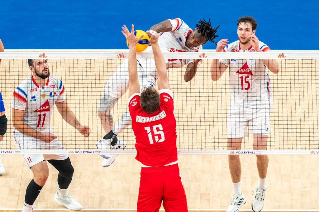 Mecz Polska - Francja w Lidze Narodów Moment meczu siatkówki, zawodnik w czerwonym stroju próbuje zablokować atak przeciwnika skaczącego nad siatką, inni zawodnicy obserwują akcję.