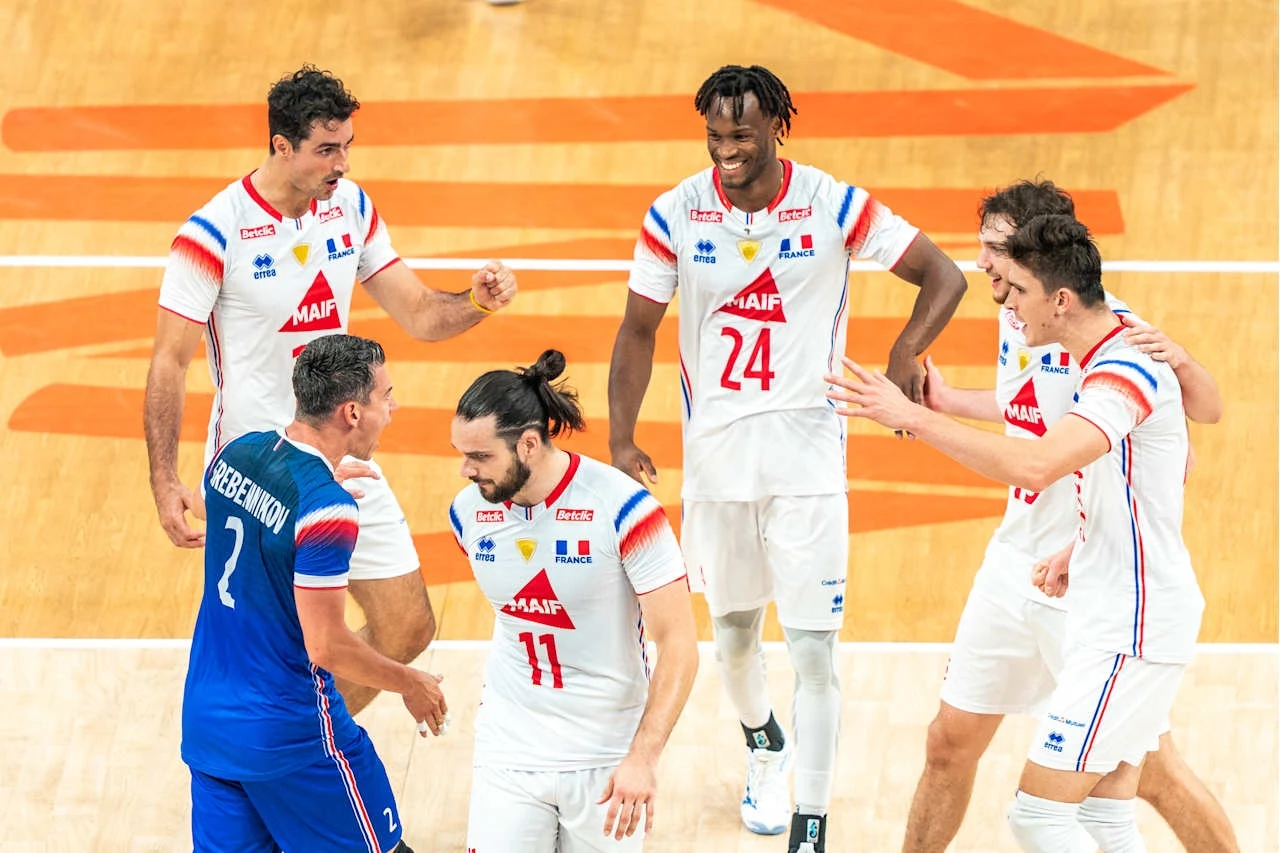 Grupa pięciu siatkarzy w białych i niebieskich strojach celebruje sukces na boisku, zawodnicy wyrażają radość i energię, wokół panuje sportowa atmosfera.