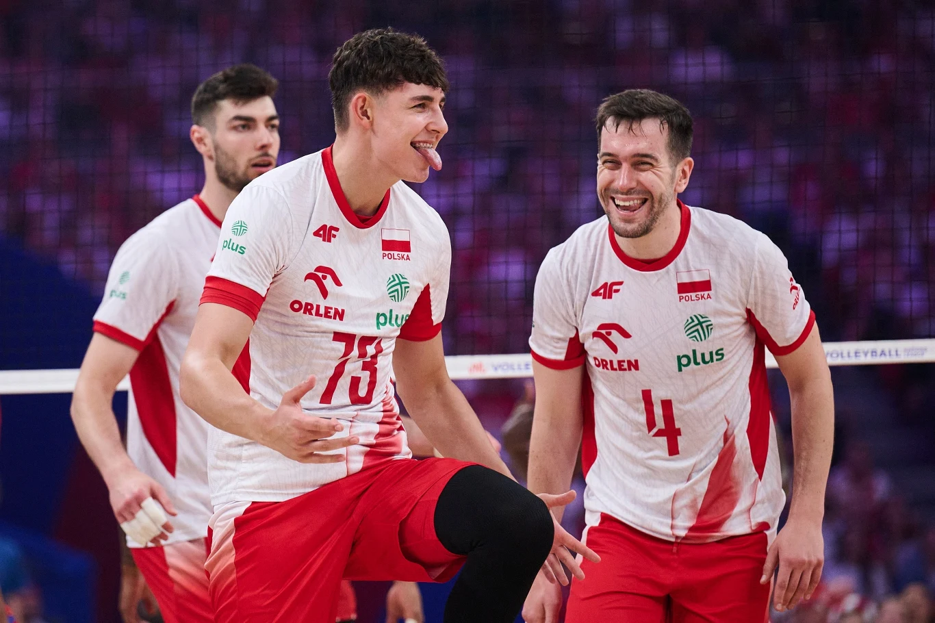 Jakub Nowak pokazał w rozmowie z nami, że nie ma kompleksów nie tylko na boisku Grupa polskich siatkarzy w białych i czerwonych strojach podczas meczu na hali sportowej, zawodnicy okazują radość i entuzjazm, jeden z nich z wystawionym językiem, a drugi szeroko się uśmiecha, w tle widoczna siatka i rozmyta publiczność.