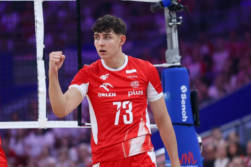 Jakub Nowak Młody siatkarz w czerwonym stroju reprezentacji Polski z zaciśniętą pięścią prezentuje sportową determinację, widoczny fragment siatki i słupka boiska, w tle rozmyta publiczność.