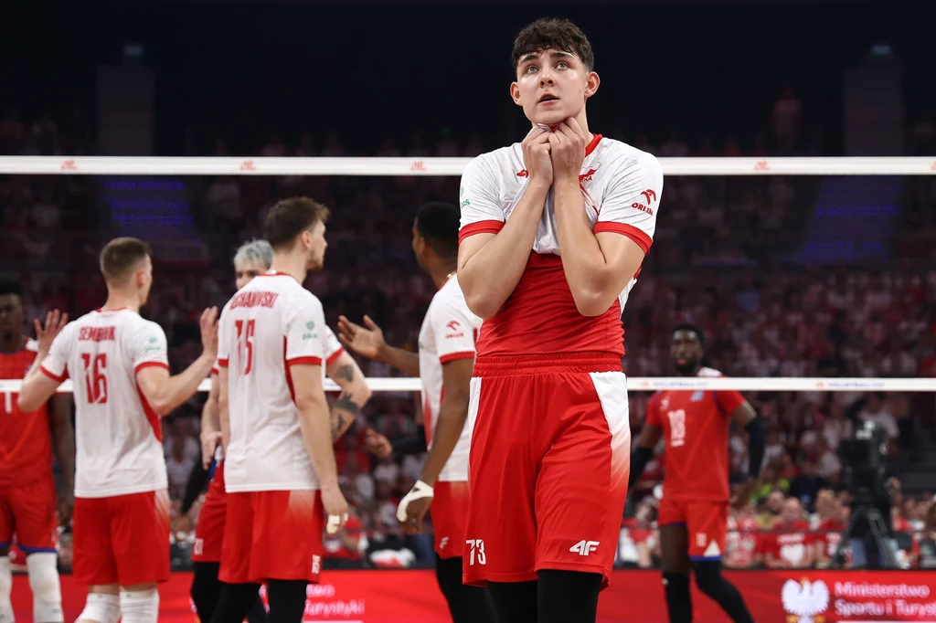 Jakub Nowak Siatkarz ubrany w czerwono-biały strój stoi na pierwszym planie na boisku siatkarskim, trzymając złożone dłonie pod brodą i wyrażając emocje. W tle pozostali zawodnicy w podobnych strojach oraz publiczność na trybunach.