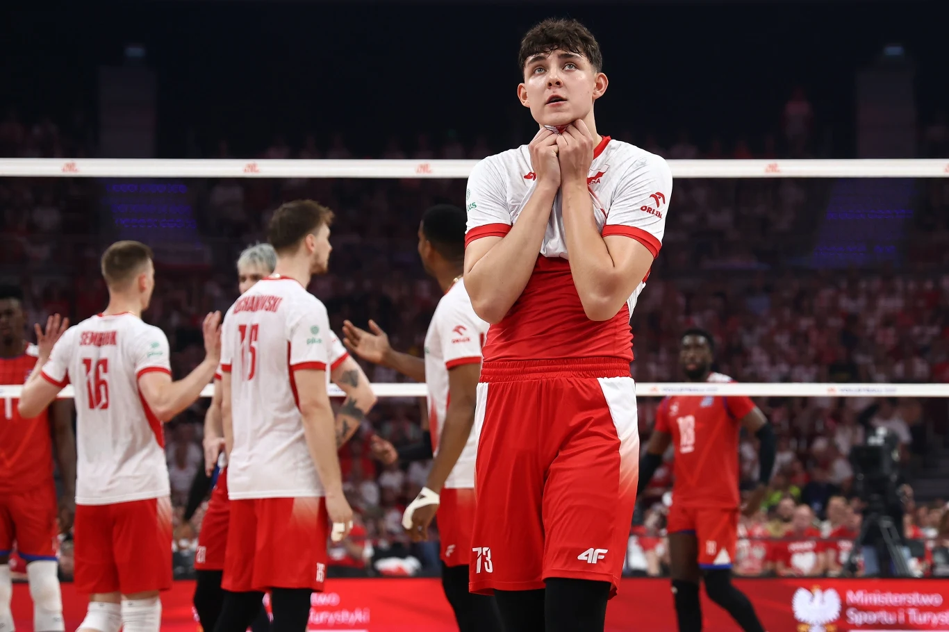 Jakub Nowak Siatkarz ubrany w czerwono-biały strój stoi na pierwszym planie na boisku siatkarskim, trzymając złożone dłonie pod brodą i wyrażając emocje. W tle pozostali zawodnicy w podobnych strojach oraz publiczność na trybunach.