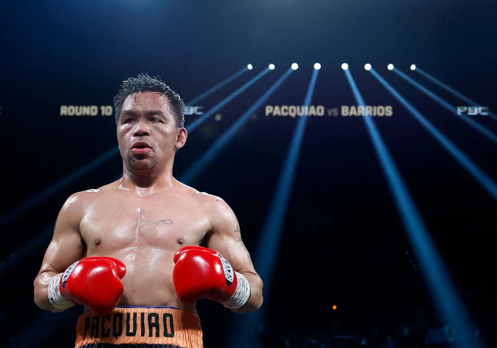 Bokser w pomarańczowych spodenkach i czerwonych rękawicach stoi na ringu, w tle reflektory skierowane na arenę oraz widoczne napisy zapowiadające walkę między Pacquiao i Barrios.