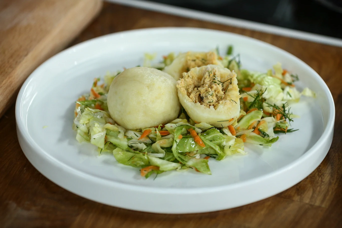 Dwa pyzy ziemniaczane, jedna przekrojona z widocznym nadzieniem, podane na sałatce z białej kapusty, marchwi oraz koperku, na białym talerzu.