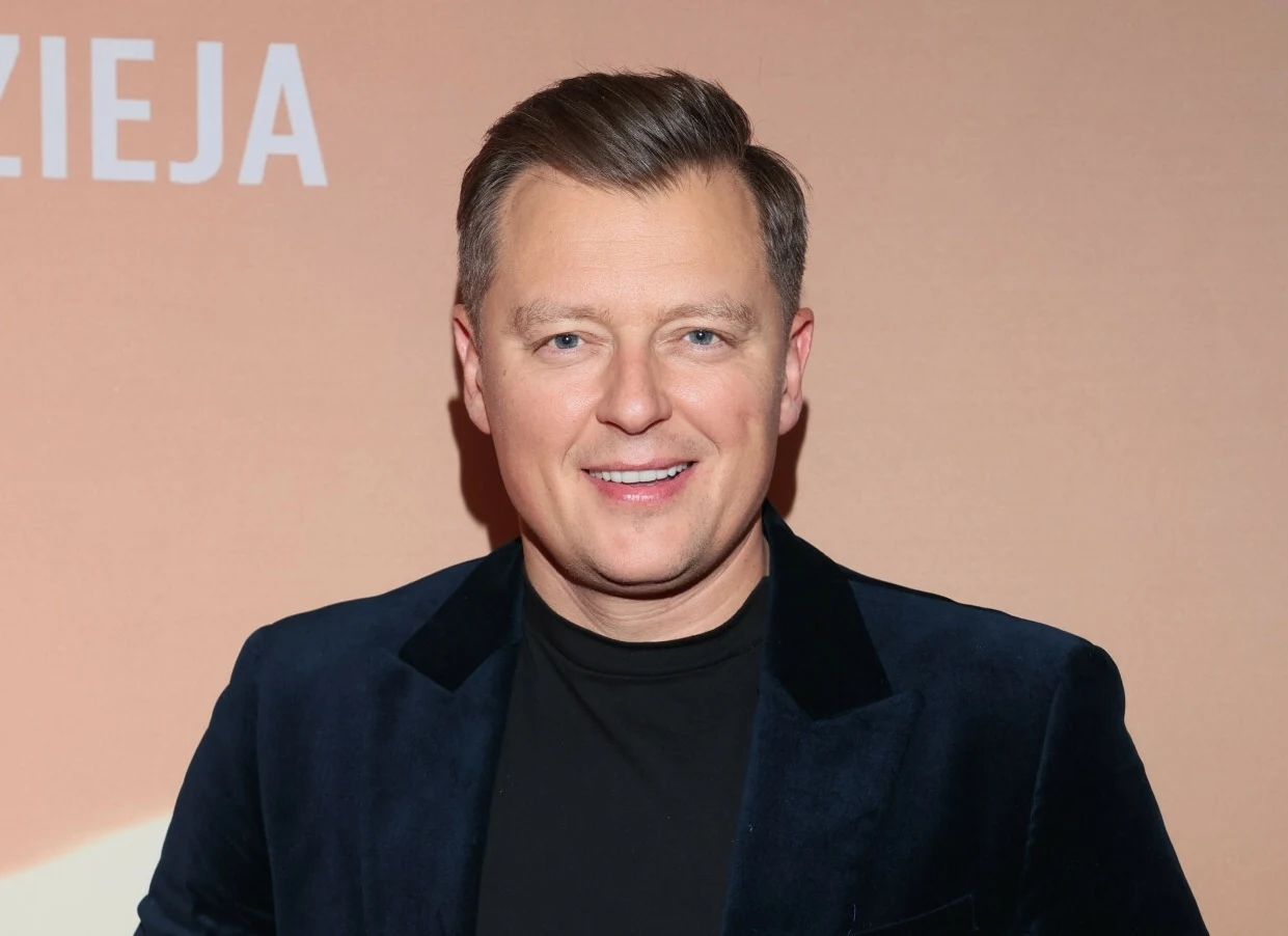 Rafał Brzozowski podał stawkę za występ na weselu Uśmiechnięty mężczyzna w ciemnej marynarce i czarnej koszulce stojący na tle jednolitej, jasnej ściany z częściowo widocznym napisem.