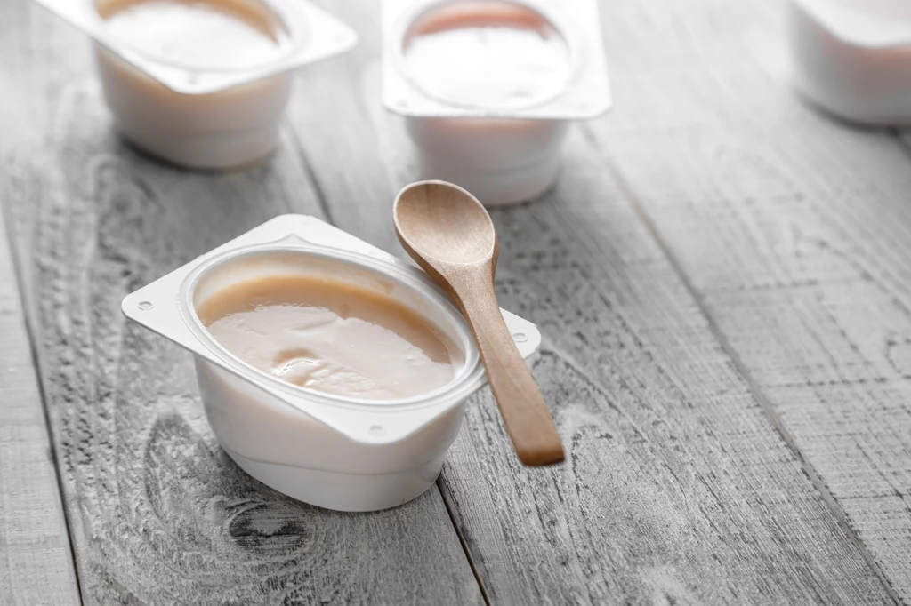 Zjadłaś serek czy jogurt w plastikowym opakowaniu? Przed wyrzuceniem przepłucz go, by pozbyć się resztek produktu Plastikowy kubeczek z kremowym deserem lub jogurtem, przykryty drewnianą łyżeczką, ustawiony na jasnym, drewnianym stole. W tle widoczne są rozmyte kolejne opakowania deseru.