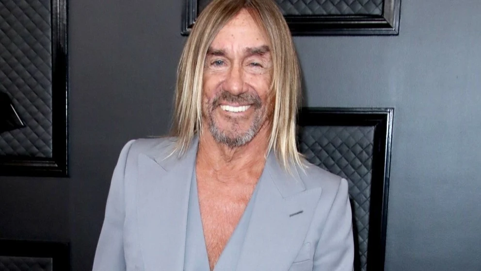 Iggy Pop Mężczyzna w średnim wieku o długich blond włosach i lekkim zaroście uśmiecha się, ubrany w szary garnitur z głębokim dekoltem, stoi na tle ciemnej ściany z geometrycznymi dekoracjami.