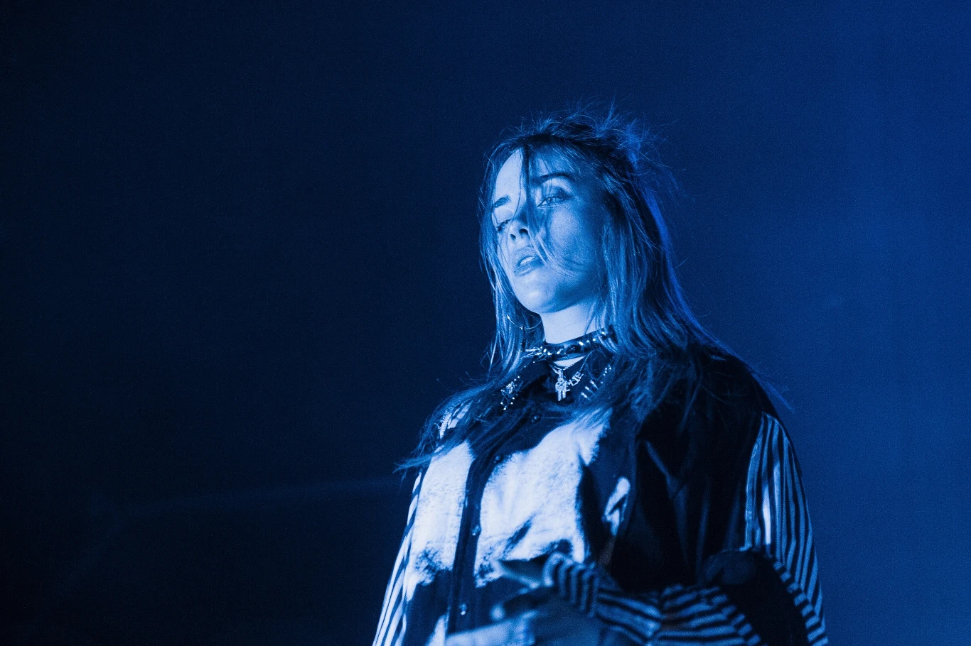 Billie Eilish zaskoczyła fanów podczas koncertu Młoda kobieta z długimi włosami ubrana w ciemną bluzę ze srebrzystymi i paskowanymi elementami, oświetlona niebieskim światłem na tle ciemnego tła.