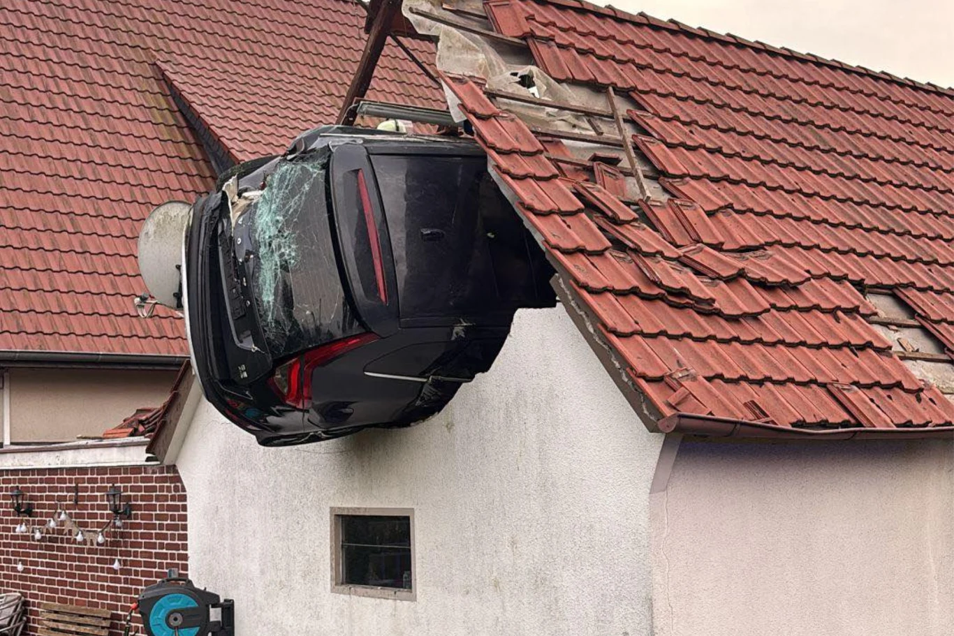 Samochód wypadł z drogi, uderzył w 7-latka i wbił się w dach. Samochód osobowy wbity w dach domu z widocznymi uszkodzeniami karoserii i zerwanymi dachówkami, ściana budynku pozbawiona fragmentu dachu, wokół brak osób.