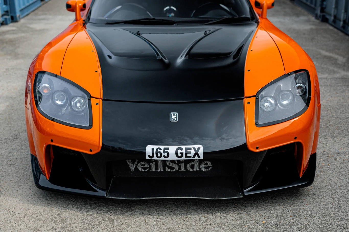 Charakterystyczny pakiet aerodynamiczny VeilSide Fortune poszerzył nadwozie RX-7 o ponad 20 cm. Czarno-pomarańczowy sportowy samochód z agresywnym body kitem VeilSide, charakterystycznymi światłami przednimi i tablicą rejestracyjną J65 GEX ustawiony na betonowej nawierzchni, skierowany przodem do oglądającego.