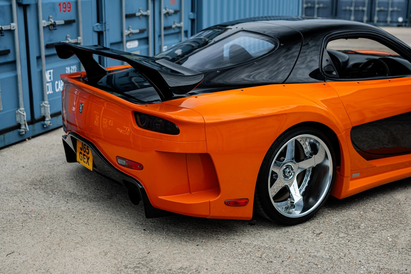Mazda RX-7 FD po renowacji i modernizacji przez warsztat RE-Amemiya, specjalizujący się w silnikach Wankla. Pomarańczowy sportowy samochód z czarnym dachem i widocznym dużym spoilerem stoi zaparkowany na tle niebieskich kontenerów przemysłowych, zwracając uwagę połyskującymi chromowanymi felgami.