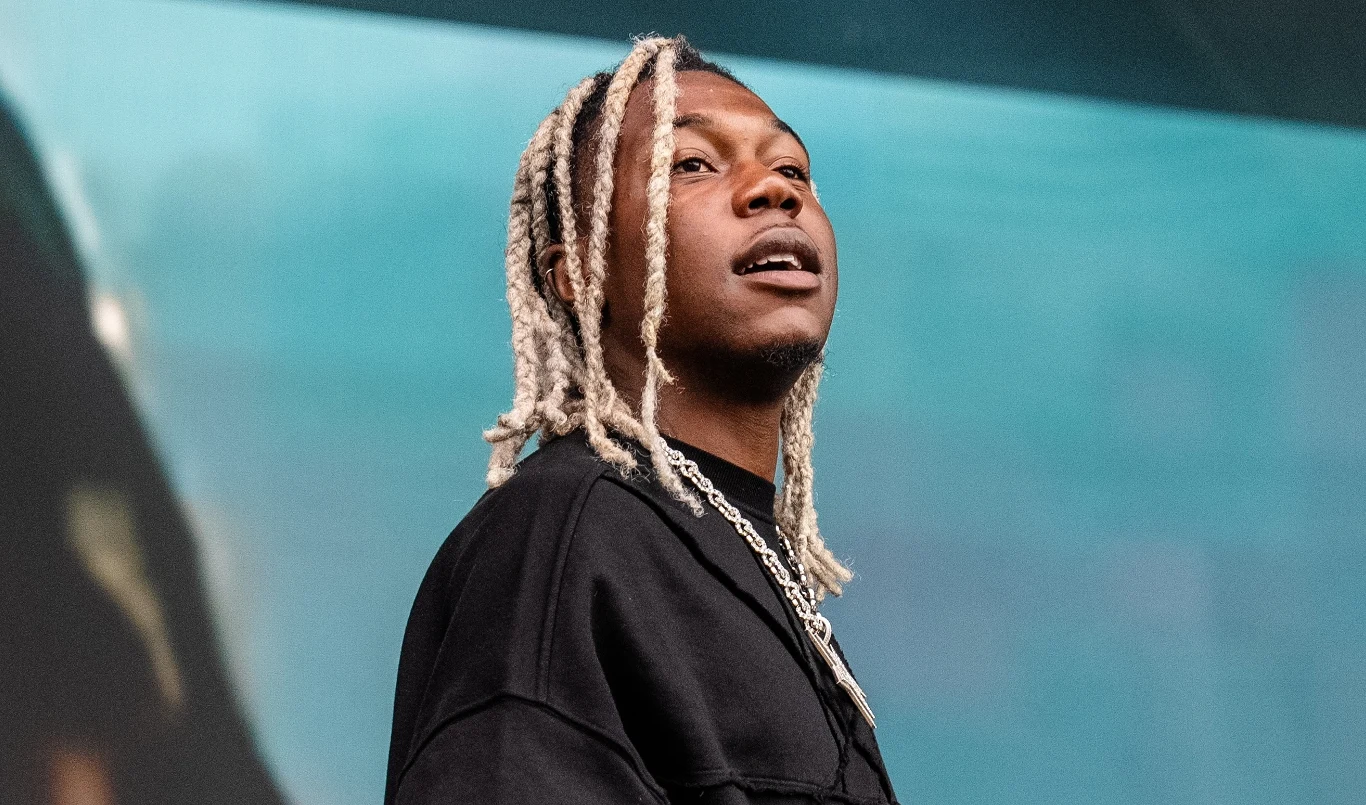 Raper Destroy Lonely zmusił nastolatka do skoku ze sceny Młody mężczyzna z jasnymi, splecionymi włosami patrzy w bok, ubrany w czarną bluzę i srebrny łańcuch na szyi, na tle rozmytego, niebieskiego tła.