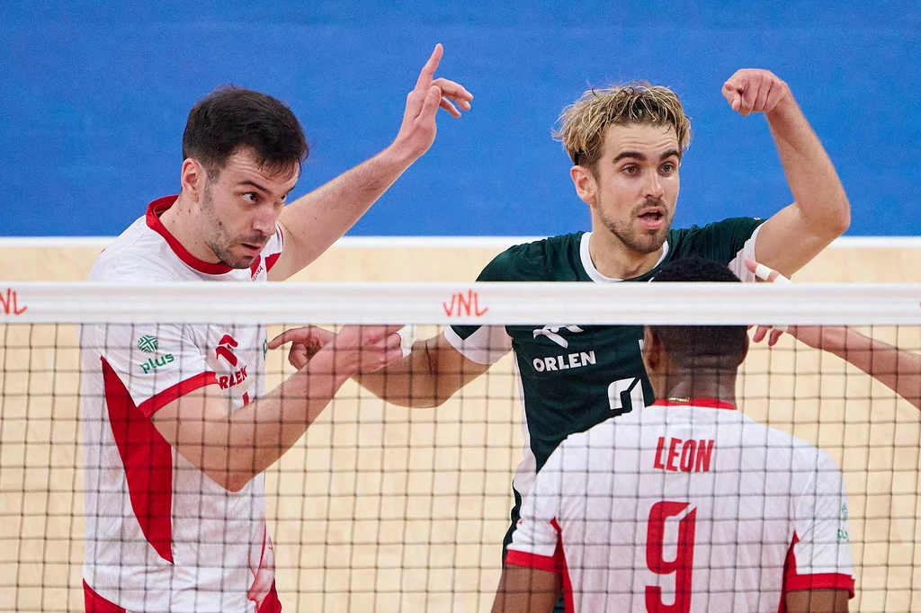 Marcin Komenda (z lewej) Trzech siatkarzy na boisku, dwóch w białych koszulkach z czerwonymi akcentami i jeden w zielonej, stoją przy siatce, gestykulując podczas rywalizacji, widoczne logo VNL na bandzie siatki.