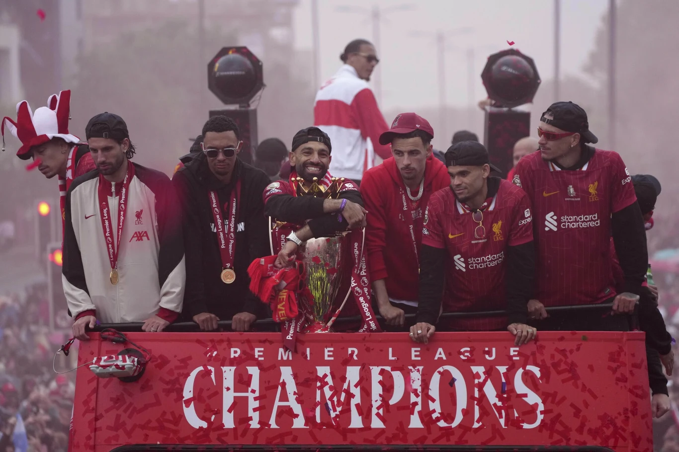 Piłkarze drużyny Liverpool w strojach klubowych świętują zdobycie mistrzostwa Premier League na otwartym autobusie przed tłumem kibiców, wraz z medalami i pucharem.