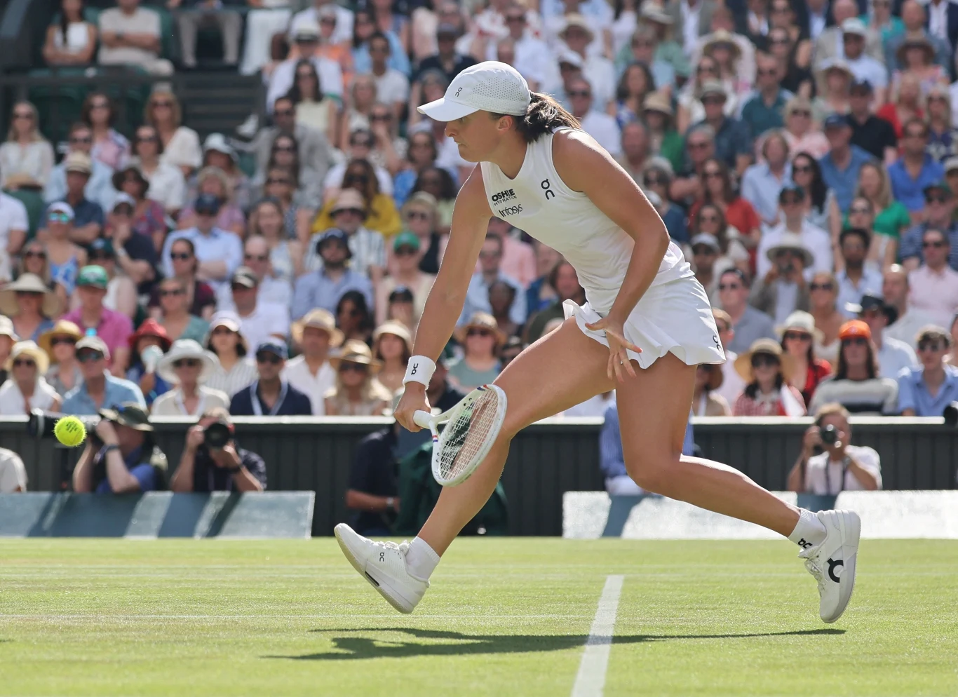 Tenisistka w białym stroju wykonuje dynamiczne uderzenie rakietą podczas meczu na trawiastym korcie, w tle widoczne trybuny pełne kibiców.