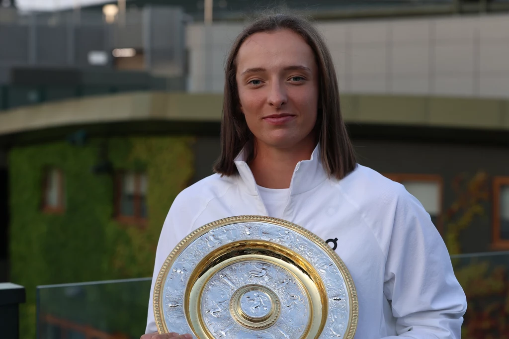 Iga Świątek Młoda kobieta ubrana w sportową białą kurtkę trzyma w rękach złoty talerzowy puchar, stojąc na tle nowoczesnego budynku pokrytego częściowo zielenią.
