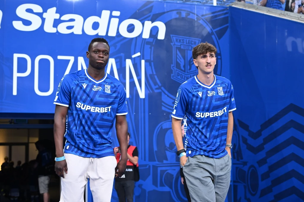 Lech Poznań jest aktywny na giełdzie transferowej Dwaj młodzi mężczyźni w niebieskich koszulkach piłkarskich klubu Lech Poznań stojący przed wejściem na stadion, na tle dużego napisu z logo klubu.
