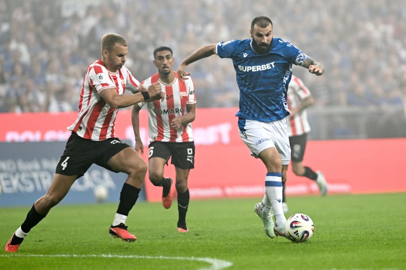 Dwóch piłkarzy w trakcie dynamicznej walki o piłkę podczas meczu, jeden ubrany w niebieską, drugi w biało-czerwoną koszulkę, w tle widoczni pozostali zawodnicy i rozmyta publiczność na stadionie.