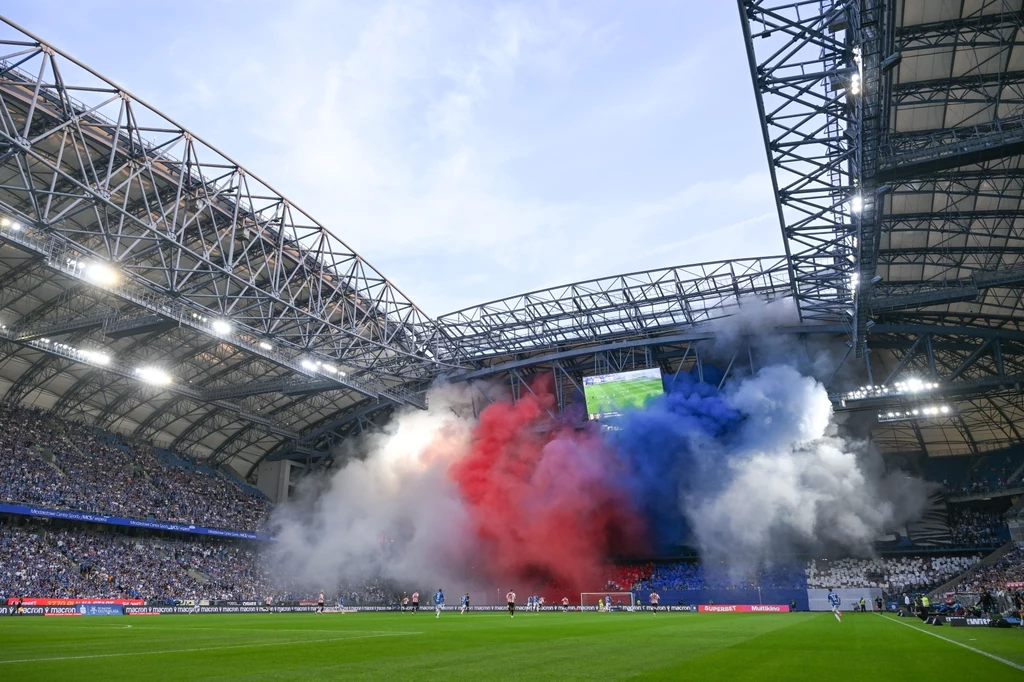 Lech Poznań marzy o Lidze Mistrzów Stadion piłkarski wypełniony kibicami, na tle trybun unosi się gęsty dym w kolorach niebieskim, białym i czerwonym, będący efektem oprawy kibicowskiej. Na boisku widoczni są piłkarze podczas trwania meczu, nad stadionem widać zamknięty dach i ekran pok...