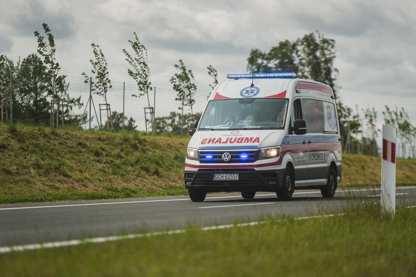 Groźna choroba znów w Polsce. Czym grozi zakażenie cholerą? Ambulans jadący z włączonymi sygnałami świetlnymi po dwupasmowej drodze otoczonej zielenią, tło stanowią rosnące drzewa i pochmurne niebo.