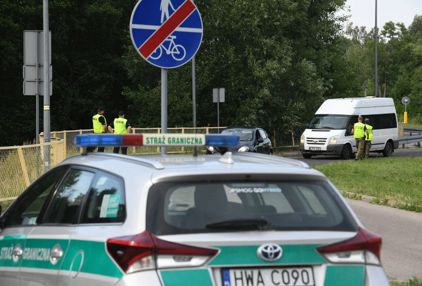 Straż Graniczna przekazała najnowsze dane dotyczące kontroli na granicach Samochód Straży Granicznej oraz białe auto dostawcze zatrzymane na poboczu, wokół którego stoją funkcjonariusze Straży Granicznej w żółtych kamizelkach odblaskowych, w tle zieleń i znaki drogowe.
