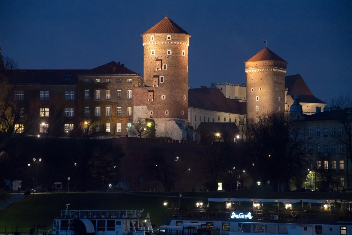 Zamek Królewski na Wawelu Oświetlony zamek na wzgórzu z charakterystycznymi ceglanymi wieżami, otoczony zabudowaniami; w dolnej części zdjęcia widoczna przystań z łodziami, całość ukazana przy zapadającym zmroku.