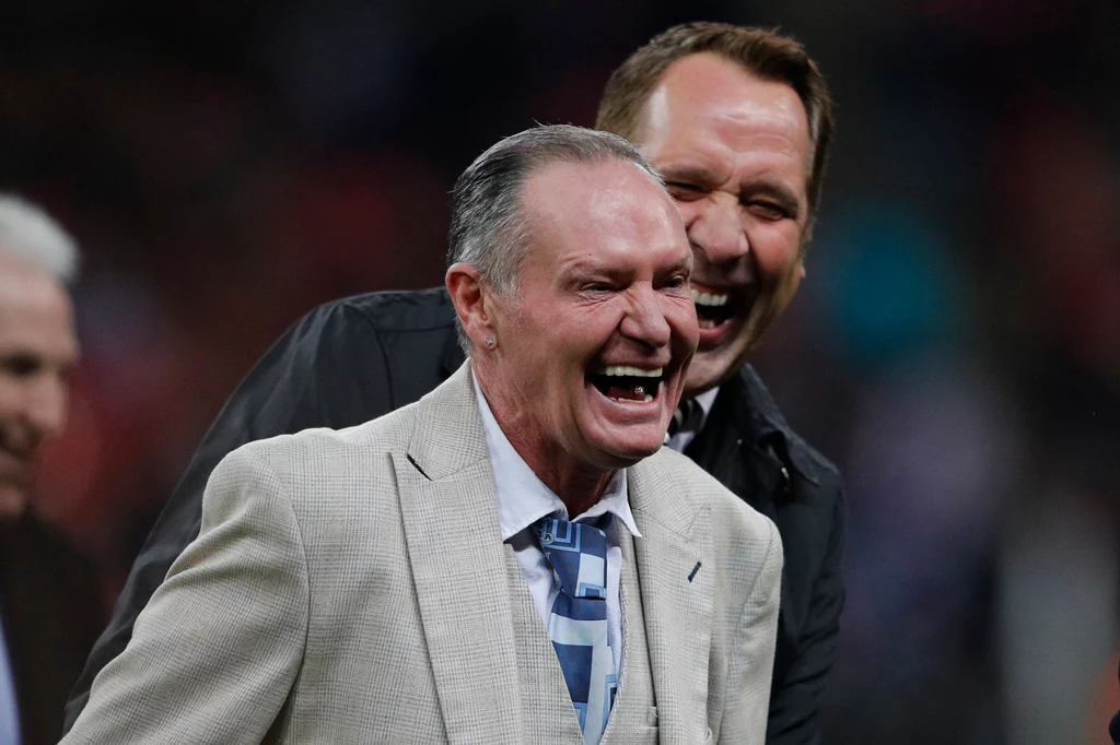 Paul Gascoigne Dwóch mężczyzn w eleganckich garniturach śmieje się radośnie, jeden z nich stoi z przodu w jasnej marynarce i krawacie w biało-niebieskie pasy, drugi w ciemnym ubraniu znajduje się za jego plecami