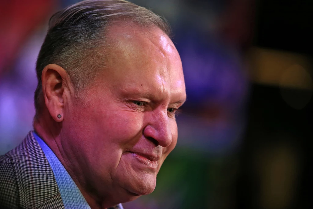 Paul Gascoigne Starszy mężczyzna z uśmiechem na twarzy, krótko ścięte włosy, kolczyk w uchu, tło rozmyte i kolorowe, skupienie na wyrazie twarzy.
