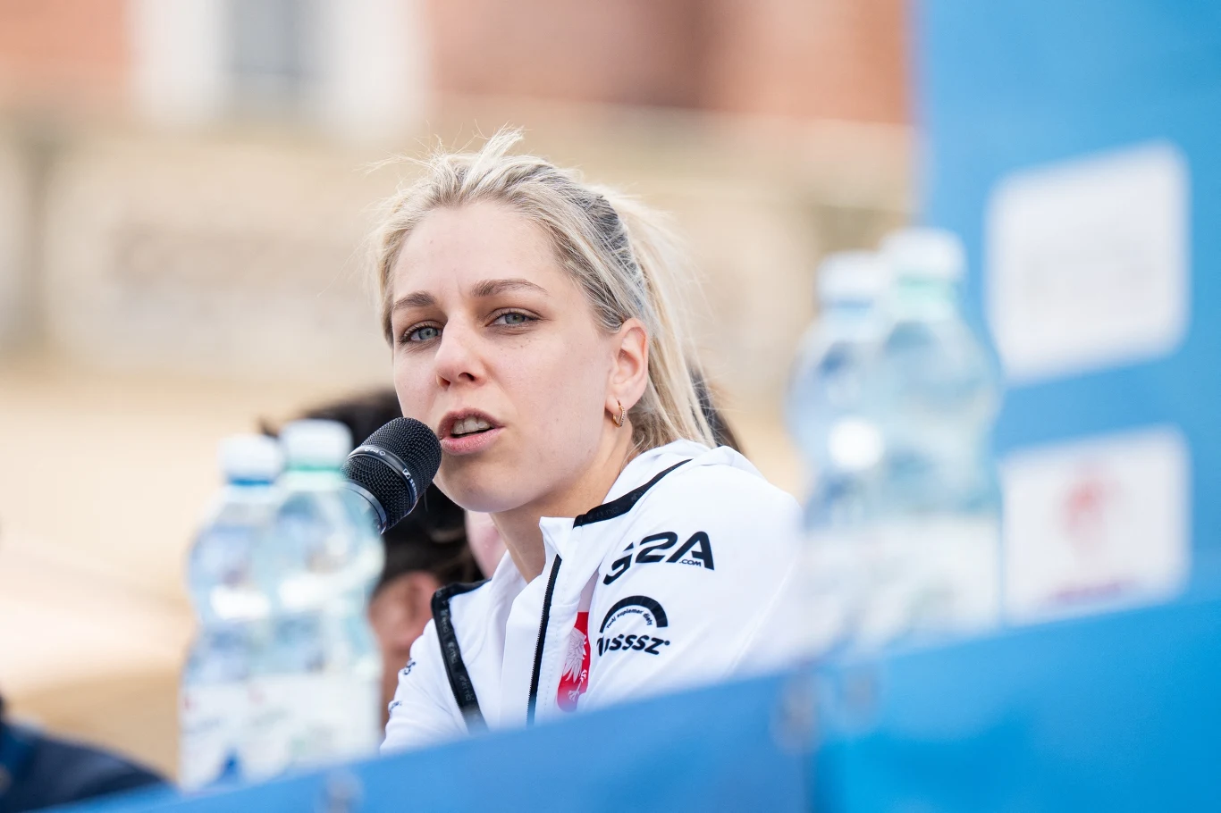 Kobieta z blond włosami mówi do mikrofonu, ubrana w sportową bluzę z logotypami, w tle rozmyty plener i kilka butelek z wodą mineralną na pierwszym planie