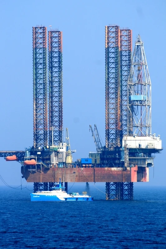 Platforma wiertnicza ustawiona na morzu z widocznymi wysokimi konstrukcjami stalowymi i żurawiami, w pobliżu znajduje się niebiesko-biały statek serwisowy