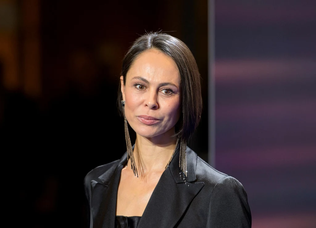 Natalia Kliczko Kobieta o krótkich, prostych włosach w eleganckiej, czarnej marynarce, stojąca na tle rozmytego, fioletowo-brązowego tła, o neutralnym wyrazie twarzy.