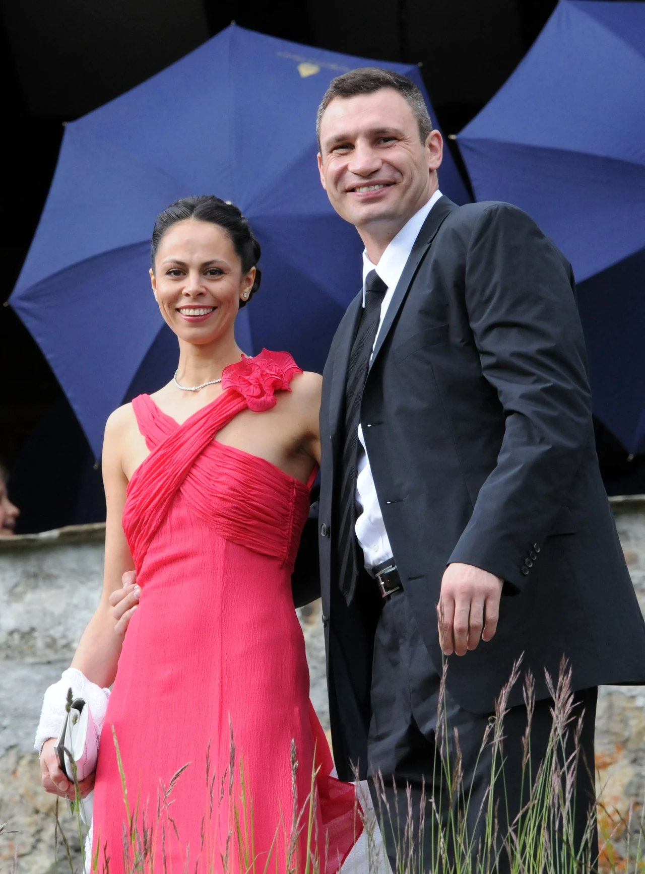 Witalij Kliczko z żoną Natalią Para ubrana elegancko, kobieta w różowej sukni z ozdobą na ramieniu, mężczyzna w garniturze z krawatem, w tle niebieskie parasole oraz fragment kamiennego muru.