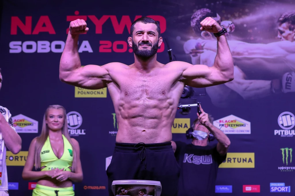 Mamed Chalidow Muskularny mężczyzna pozuje z uniesionymi ramionami na scenie podczas oficjalnego ważenia zawodników, w tle stoją kobieta w stroju promocyjnym oraz inni członkowie obsługi wydarzenia.