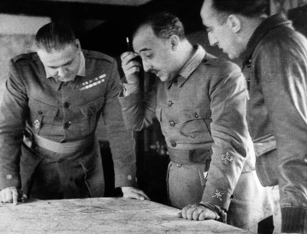 Francisco Franco (w środku) podczas Hiszpańskiej Wojny Domowej w 1936 roku Trzech mężczyzn w mundurach wojskowych pochylonych nad dużą mapą, prowadzących poważną analizę lub dyskusję, światło skupia uwagę na nich, a mimika sugeruje koncentrację i zaangażowanie.