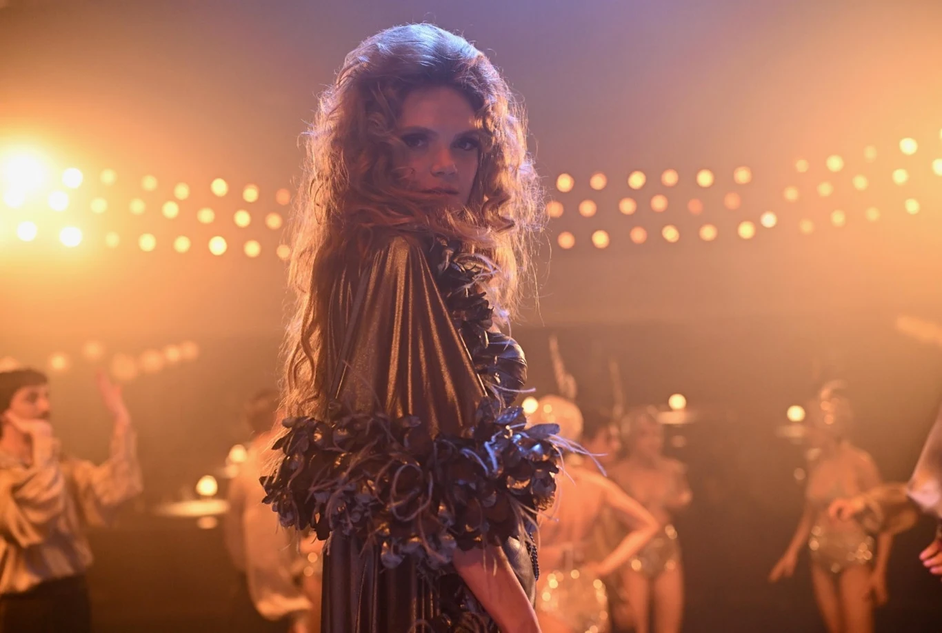 Kobieta w efektownym, błyszczącym stroju ze zdobieniami z piór stoi na środku sceny, skierowana twarzą do obiektywu, wokół niej inne osoby w tanecznych pozach, rozświetlone światłami w klimacie kabaretu.