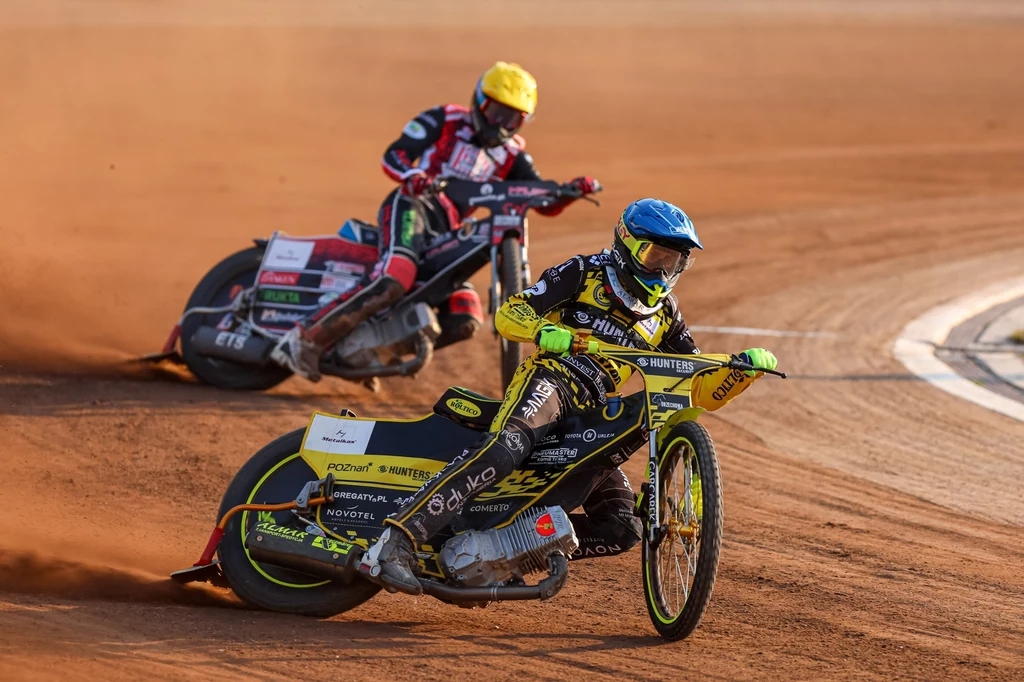 Dwóch żużlowców w pełnym pędzie podczas wyścigu na torze, motocykle unoszące kurz z ziemistej nawierzchni, sportowe kombinezony i kaski w jaskrawych kolorach, dynamiczna scena sportowej rywalizacji.