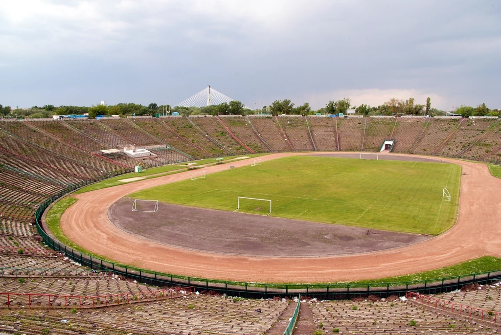 Stadion Dziesięciolecia Opuszczony stadion z pustymi trybunami, zarośniętym boiskiem piłkarskim i bieżnią ziemną otaczającą murawę, w tle widoczne drzewa oraz fragment nowoczesnego mostu.