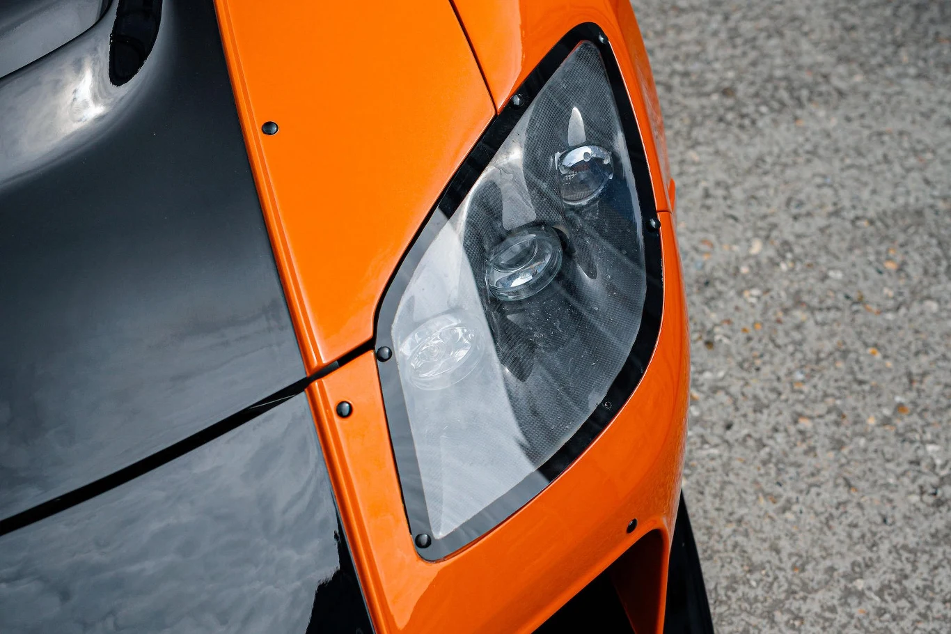 Mazda RX-7 FD VeilSide, filmowy samochód Hana z "Tokio Drift”, sprzedany za rekordową kwotę. Zbliżenie na przedni reflektor sportowego samochodu w kolorze pomarańczowo-czarnym, wyraźnie widoczne detale obudowy lampy oraz fragment karoserii pojazdu na tle asfaltowej nawierzchni.