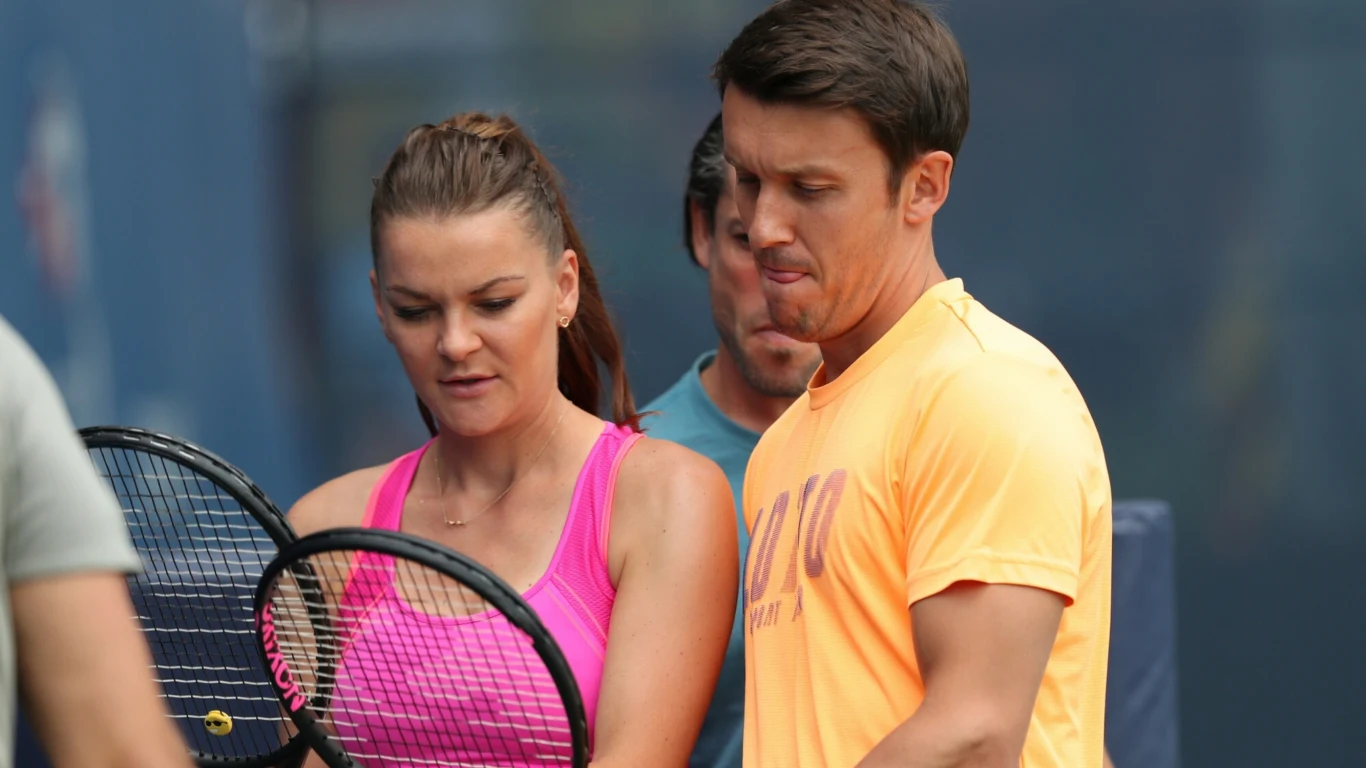 Agnieszka Radwańska i Dawid Celt Dwie osoby ubrane sportowo trzymają rakiety tenisowe i koncentrują się na wspólnej czynności, tło rozmyte w odcieniach niebieskich, scena ma charakter treningowy.