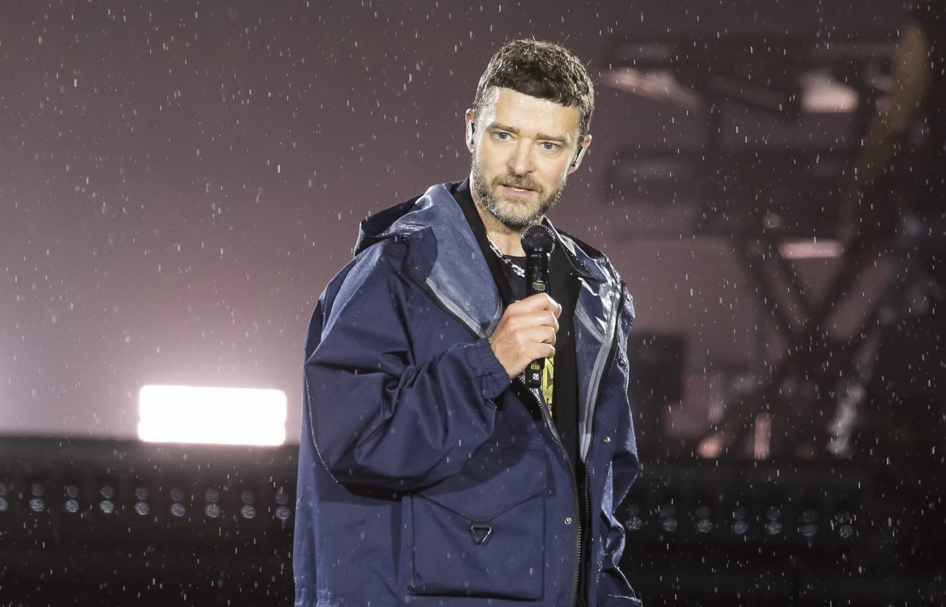 Justin Timberlake rozczarował swoich fanów na koncercie Mężczyzna w kurtce przeciwdeszczowej trzyma mikrofon, stoi na scenie podczas deszczu, w tle rozmyte światło i konstrukcja sceniczna.