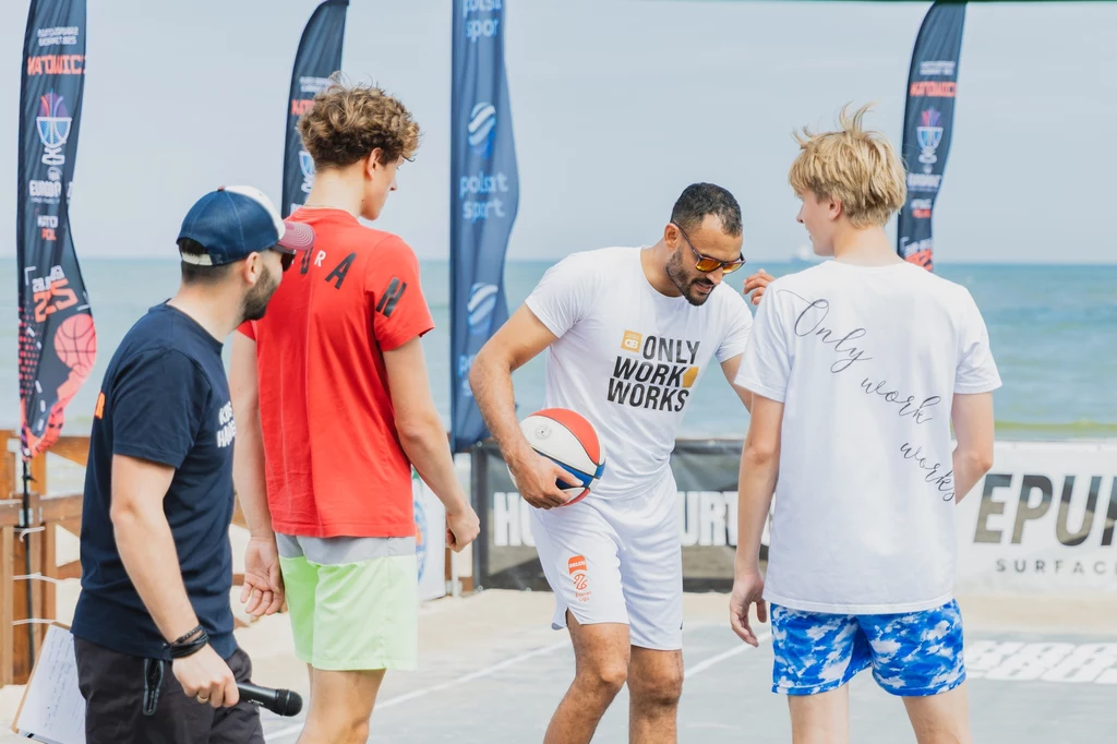 Grupa młodych mężczyzn na boisku do koszykówki przy plaży, jeden z nich trzyma piłkę i nosi sportowy strój z napisem Only Work Works, w tle morze oraz flagi promujące wydarzenie sportowe.