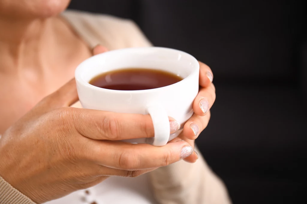 Osobom z podwyższonym cholesterolem zaleca się picie herbaty - zarówno zielonej jak i czarnej Dłonie osoby trzymającej dużą białą filiżankę z gorącą herbatą, w tle rozmyte ciemne tło podkreśla relaksacyjny charakter chwili.