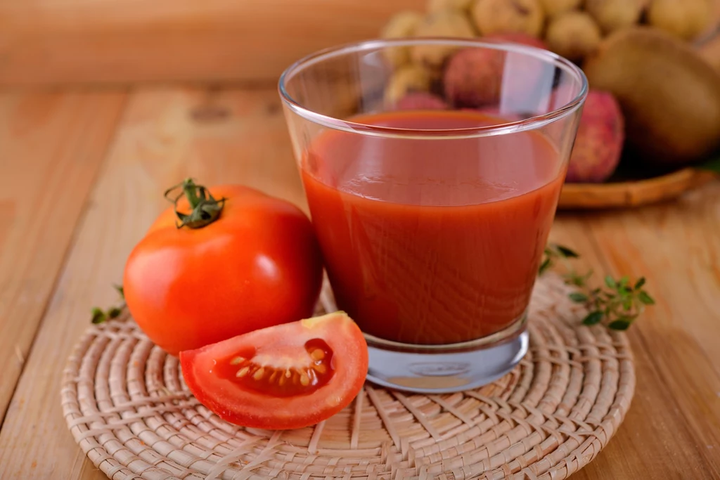 W zbijaniu cholesterolu pomocny będzie też sok pomidorowy Szklanka soku pomidorowego ustawiona na wiklinowej podkładce, obok znajduje się dojrzały pomidor oraz przekrojony kawałek pomidora, w tle widać rozmyte owoce.