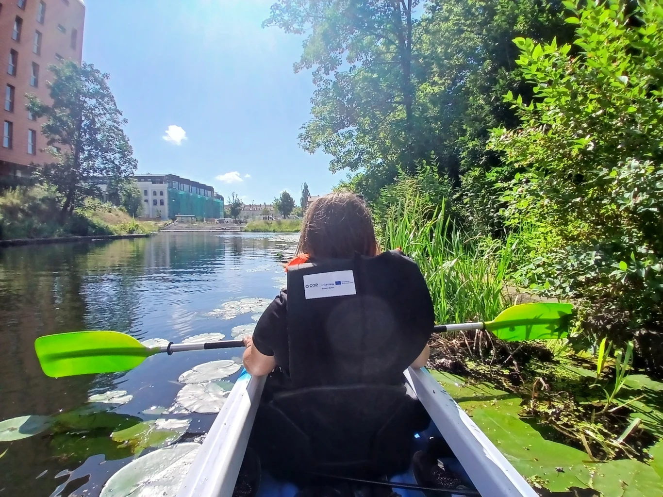 EkoKajak w Gdańsku - fot. Gdański Ośrodek Sportu Osoba z wiosłem siedzi w kajaku płynącym po spokojnym kanale, otoczonym zielenią i roślinami wodnymi, po lewej w tle widoczne nowoczesne budynki, a słońce świeci z niemal bezchmurnego nieba.