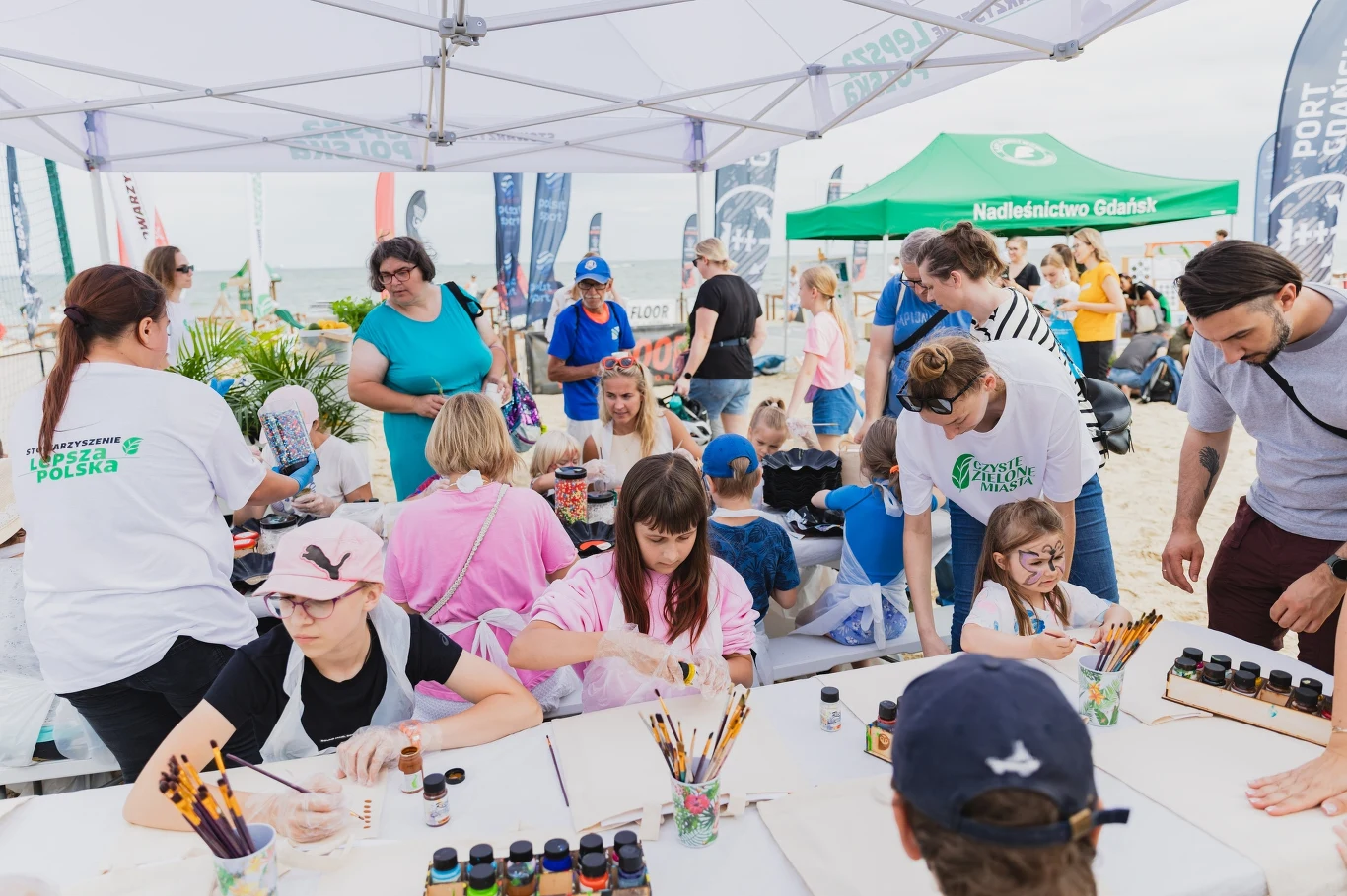 Zajęcia przy stoisku Stowarzyszenia - fot. UM Gdańsk Warsztaty artystyczne na świeżym powietrzu, grupa dzieci i dorosłych maluje przy stołach pod namiotem na plaży, liczne farby, pędzle i kolorowe akcesoria, w tle namioty organizacyjne oraz uczestnicy wydarzenia.