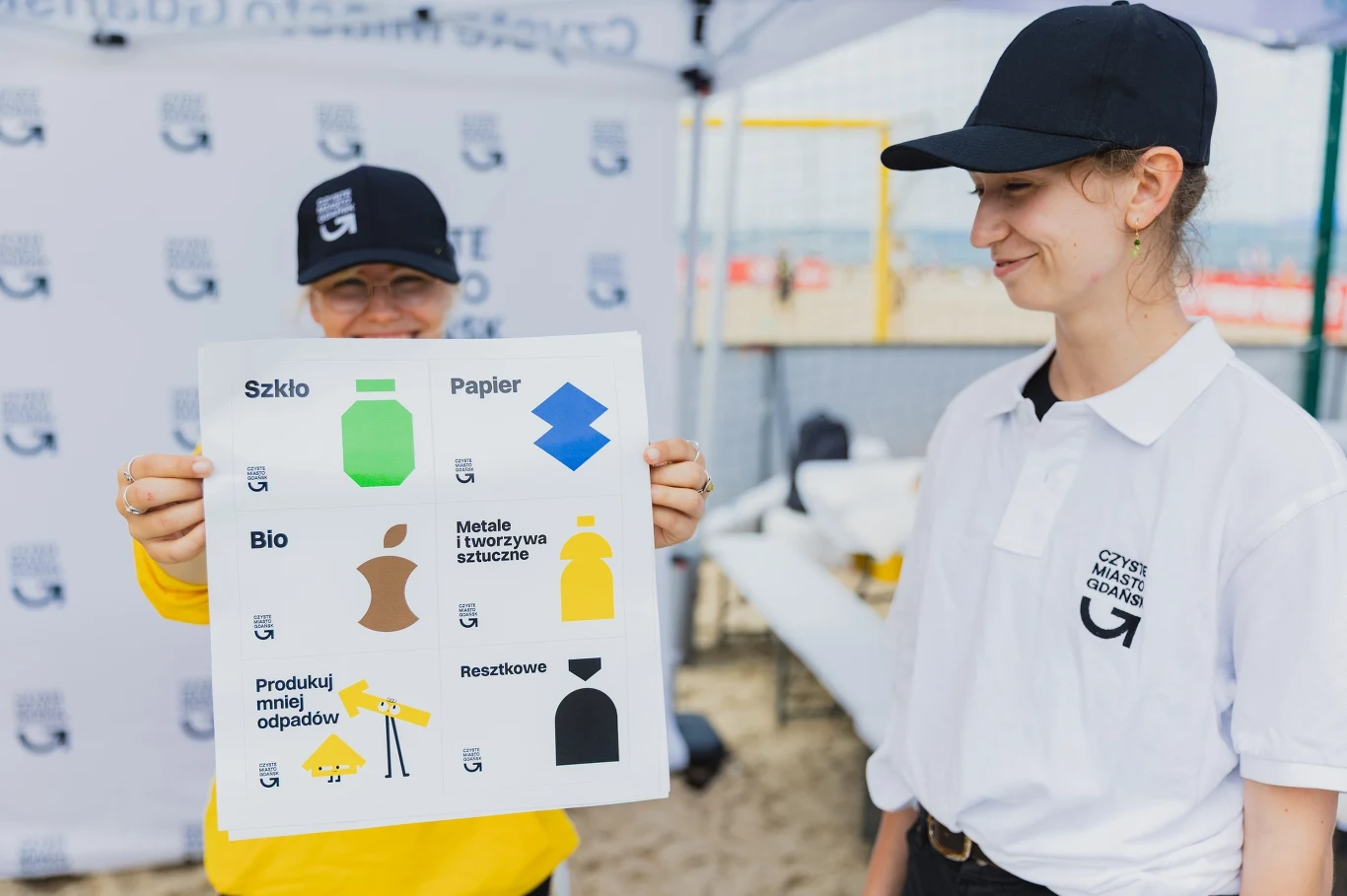Zajęcia z segregacji odpadów - fot. UM Gdańsk Dwie osoby w ubraniach z logo akcji stoją pod namiotem na plaży, jedna trzyma tablicę edukacyjną dotyczącą segregowania śmieci z kolorowymi symbolami różnych frakcji odpadów.